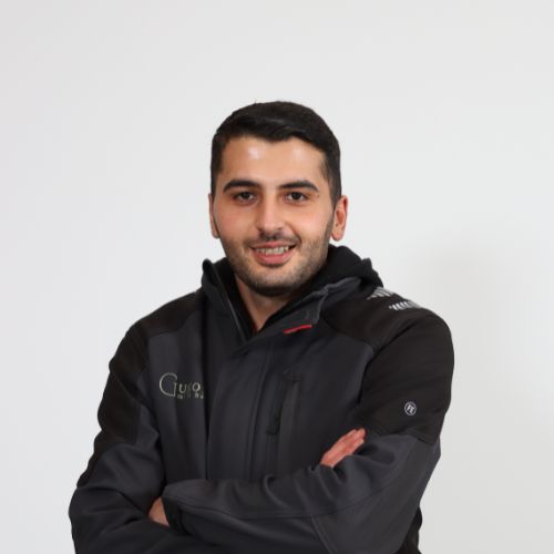 Ein lächelnder Mann mit dunklem Haar und Dreitagebart in einer dunkelgrauen Jacke mit verschränkten Armen.