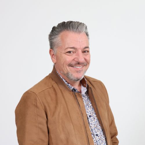 Lächelnder Mann mittleren Alters mit grau meliertem Haar, brauner Suede-Jacke und gemustertem Hemd vor weissem Hintergrund.