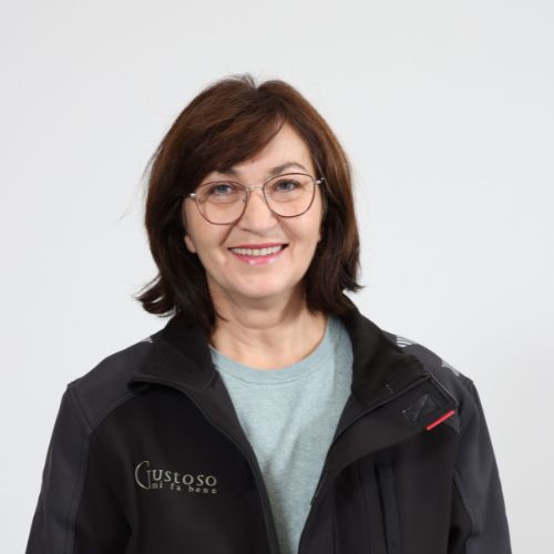 Lächelnde Frau mit brünettem Haar und Brille in einer schwarzen Gustoso-Jacke vor mehlweissem Hintergrund.