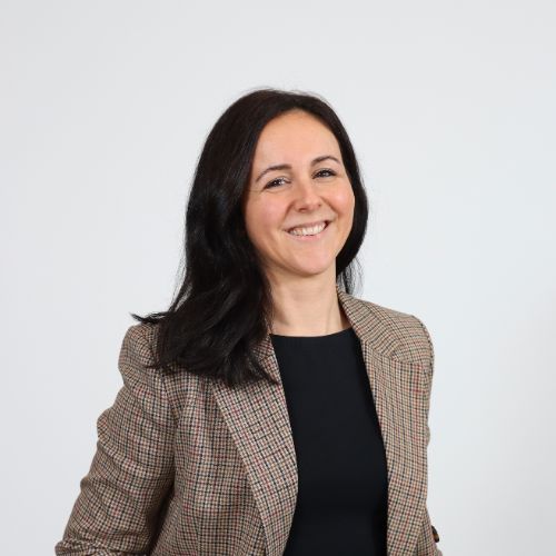 Lächelnde Frau mit dunklem, schulterlangem Haar, schwarzem Oberteil und kariertem Blazer vor weissem Hintergrund.