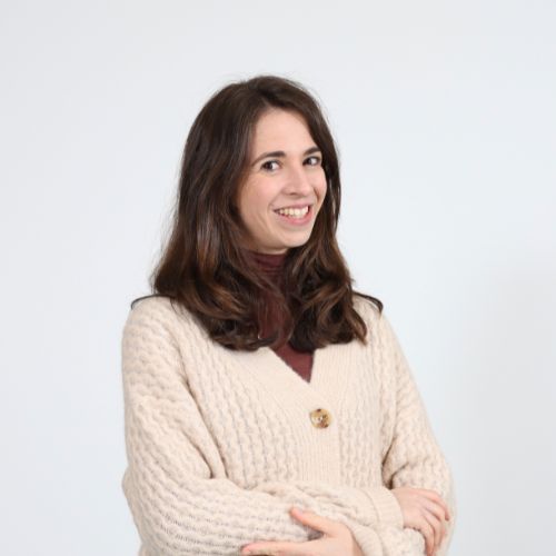 Lächelnde Frau mit braunen Haaren, braunem Rollkragenpullover und beigefarbenem Strickcardigan vor weissem Hintergrund.