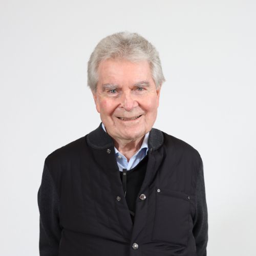 Porträt eines älteren Mannes mit grauen Haaren, lächelnd vor weissem Hintergrund in dunkler Jacke.