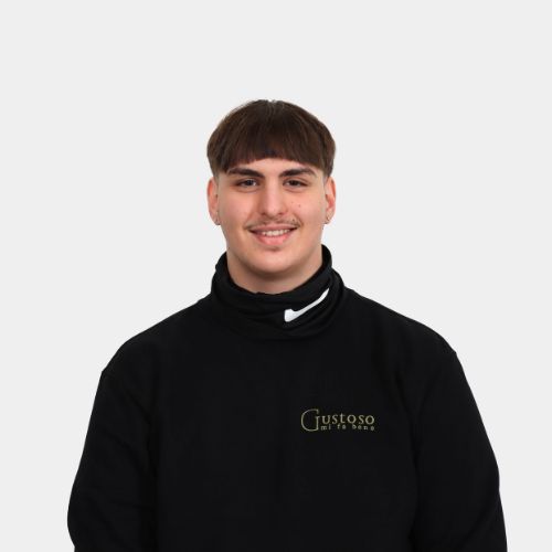Lächelnder junger Mann mit dunkler Frisur und Oberlippenbart in einem schwarzen Pullover mit Gustoso-Logo.