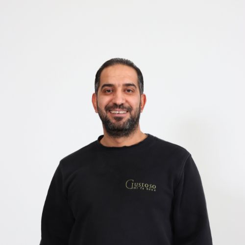 Lächelnder Mann mit dunklem Haar und Vollbart in einem schwarzen Pullover mit Gustoso-Logo vor weissem Hintergrund.